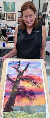 Woman holding up her watercolor painting of a tree and a sunset.